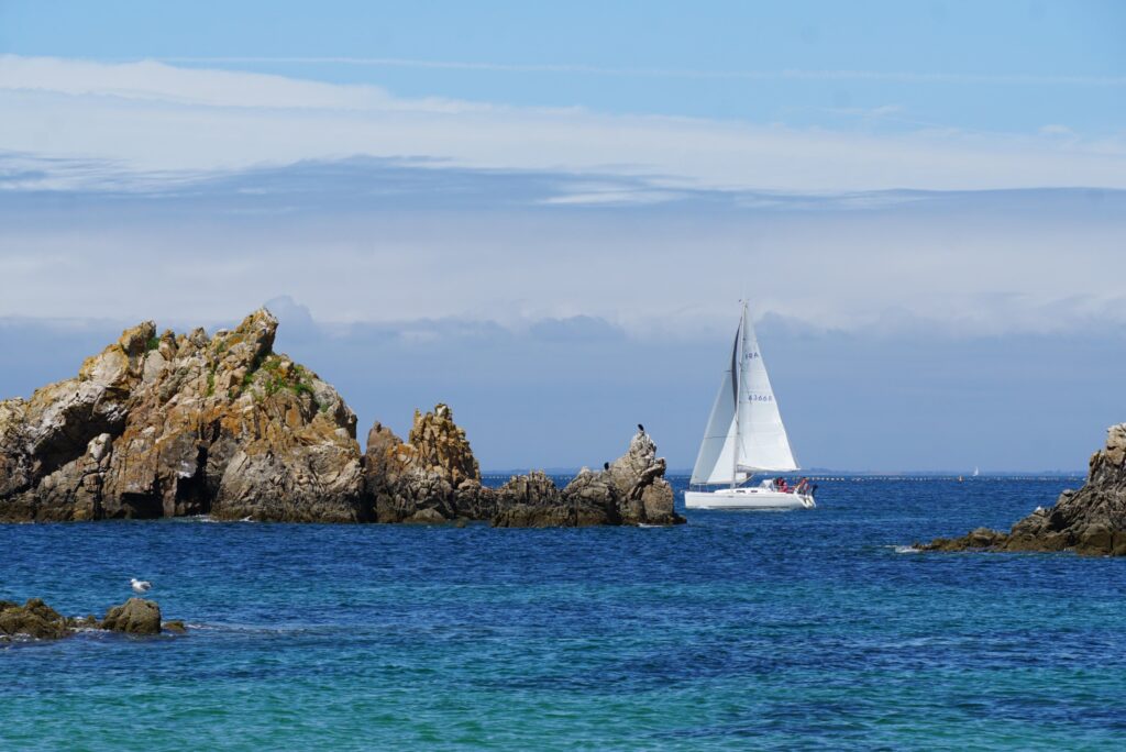 Eine Yacht mit weißen Segeln segelt auf dem offenen Meer, im Vordergrund einige Felsformationen.