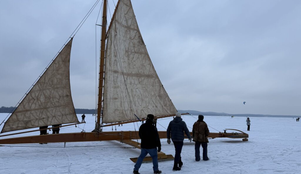 Der Himmel ist grau, der See schneebedeckt, im Vordergrund steht eine große Eisyacht aus Holz.
