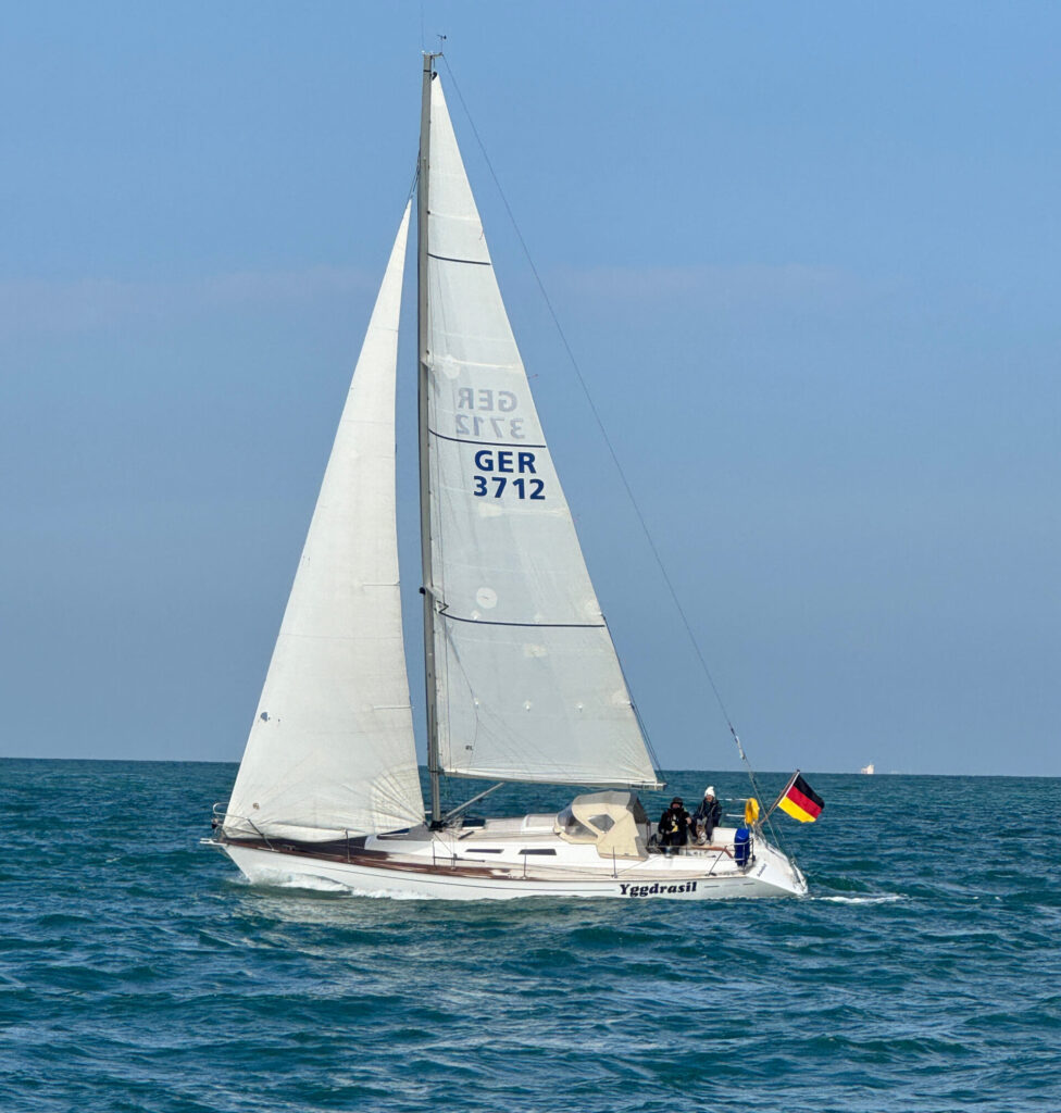 Eine Yacht segelt bei viel Wind, die Menschen im Cockpit sind dick angezogen. Am Heck weht die deutsche Flagge.