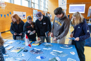 Junge Teilnehmende des Jugendseglertreffens stehen vor einem Tisch mit Informationsmaterialien und erkunden die verschiedenen angebotenen Programme.