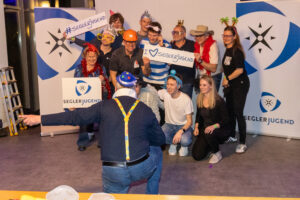 Teilnehmende des Jugendseglertreffens stehen vor mehreren Aufstellern und halten drei Schilder mit der Aufschrift „Seglerjugend“. Sie tragen witzige Kopfbedeckungen, während der Fotograf im Vordergrund ihnen zeigt, wohin sie schauen sollen.