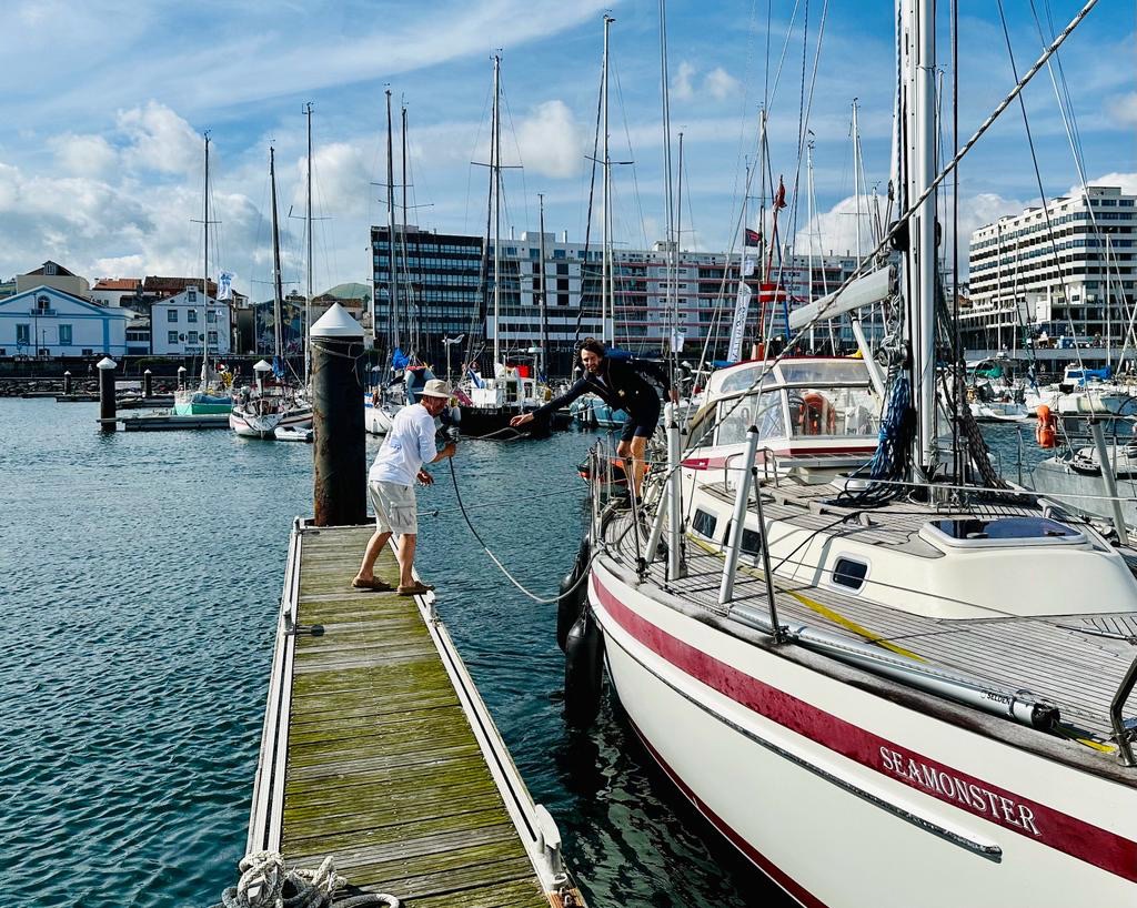 Ein Segler auf einem Boot reicht einem Mann auf einem Steg ein Seil - er will am Steg in einem Yachthafen festmachen.