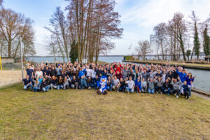 Gruppenfoto der Teilnehmenden des Jugendseglertages: Die Gruppe steht auf einer Wiese am Wasser, in der Mitte das Maskottchen der Seglerjugend „Skippi“.