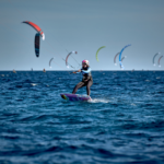 Eine Kitesurferin lässt sich von ihrem roten Drachen durch Wasser ziehen. Im Hintergrund gibt es noch viele weitere Kiter, ihre Kites sind am Himmel zu sehen.