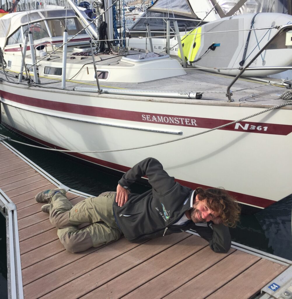 Ein Mann liegt auf einem Steg vor seiner Segelyacht und freut sich.