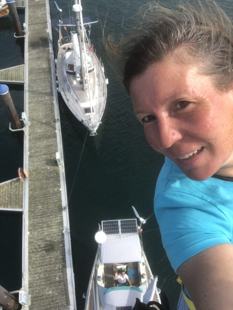 Eine Frau ist ganz oben im Mast ihrer Sgeleyacht und macht von dort aus ein Foto nach unten - dort steht eine andere Frau im Cockpit und winkt nach oben.
