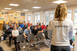 Teilnehmende des Jugendseglertreffens sitzen in einem Seminarraum und hören dem Vortrag des Juniorteams der Seglerjugend zu. Rechts im Bild steht ein Mitglied des Juniorteams in einem weißen T-Shirt mit der Aufschrift „Juniorteam“