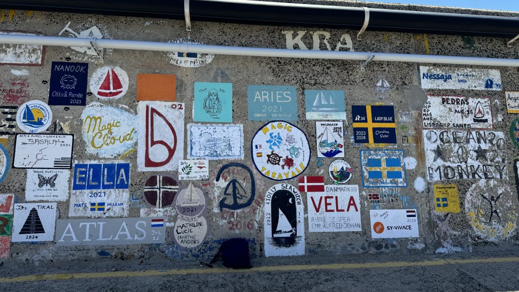 An einer grauen Hafenmauer hängen ganz viele Bilder - sie sind alle Logos von Segelyachten.