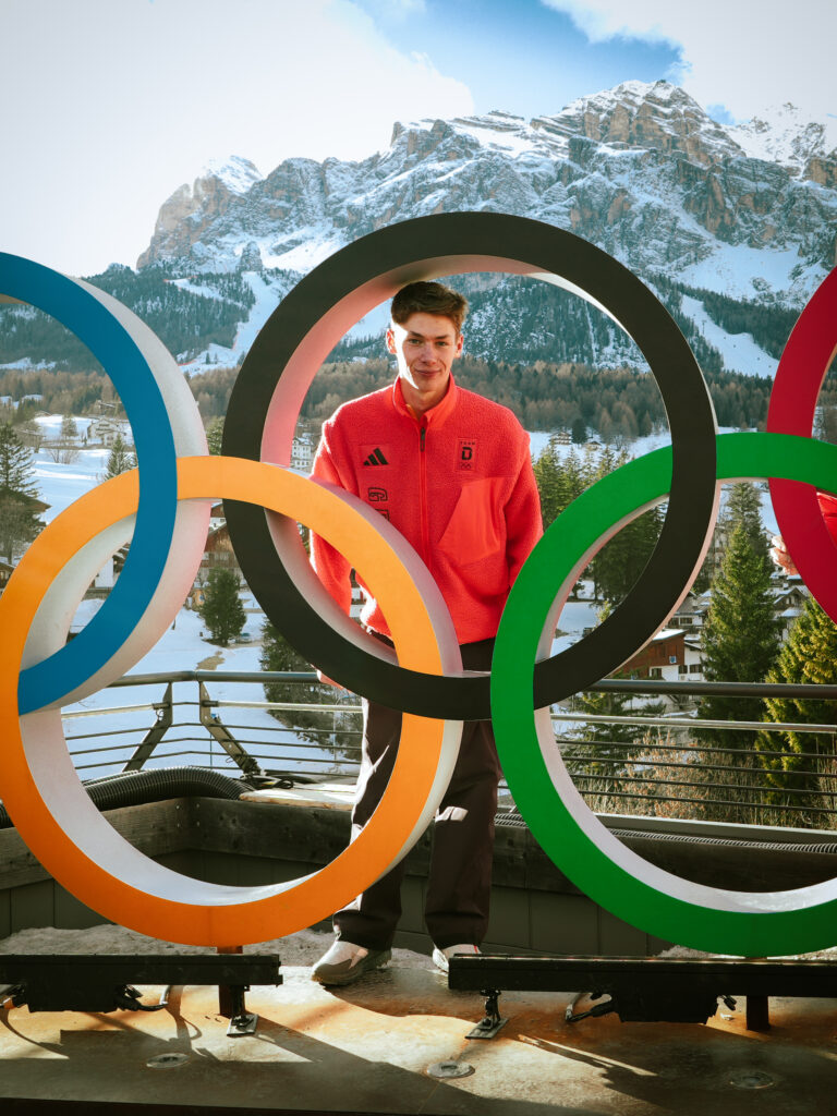 Auf dem Foto steht Patrick Schulz, Teilnehmer des Deutschen Olympischen Jugendlagers in Milano Cortina, zusammen mit den Olympischen Ringen vor einer Bergkulisse.