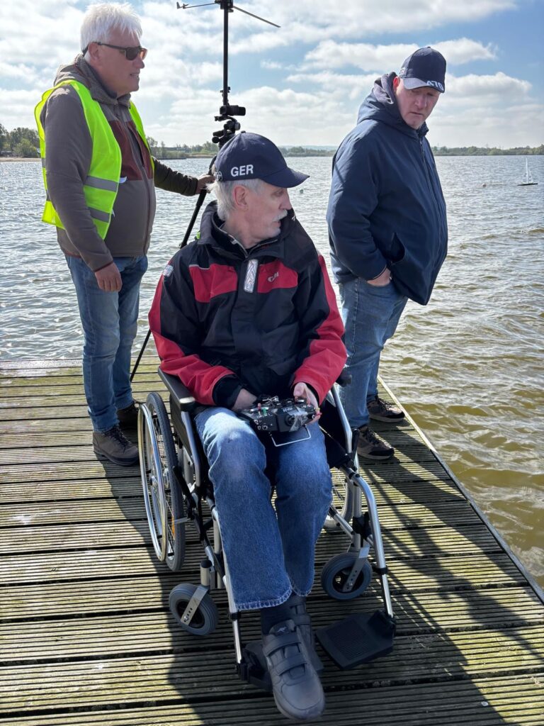 Ein Mann sitzt in einem Rollstuhl auf einem Steg. Er steuert über eine Fernsteuerung eine Modell-Segelyacht auf dem Wasser.