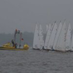Startende Jollen mit weißen Segeln in denen ein O ist auf einem See neben einem gelben Motorboot. Das Wetter ist schlecht, es ist grau und regnet.