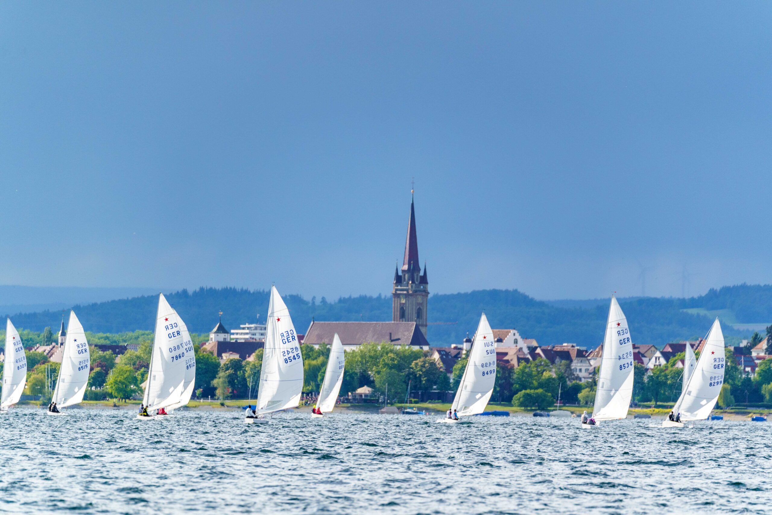 Die Kulisse von Radolfzell am Bodensee, im Vordergrund mehrere Segelboote mit weißen Segeln und einem Stern im Segel.