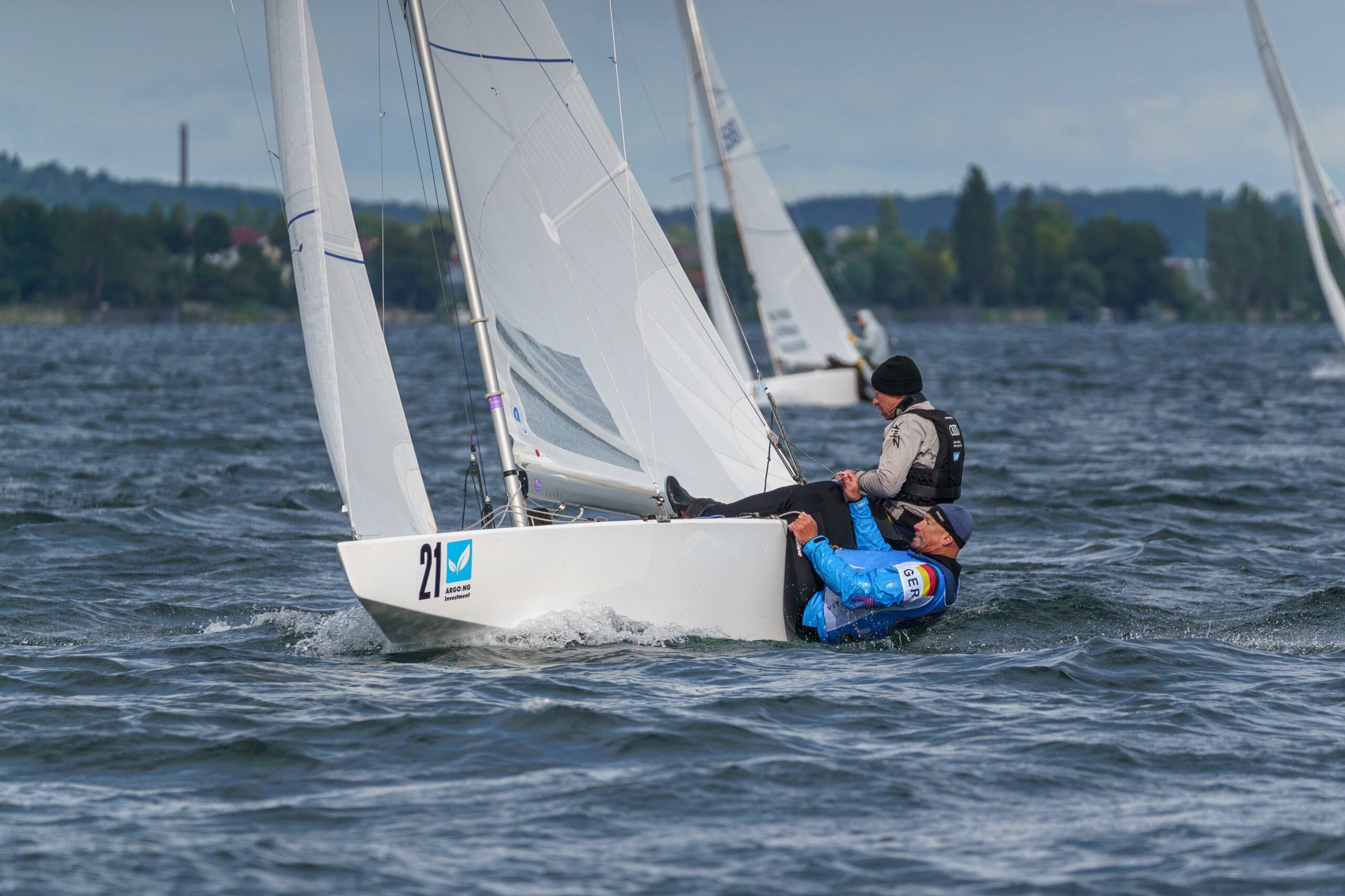 Zwei Männer segeln auf einem weißen Kielboot, der Vordere hängt mir rechtwinklig gebeugten Beinen an der Außenkante.