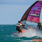 Ein Windsurfer rast durchs Wasser. Gischt spritzt hoch. Im Hintergrund beobachten Menschen auf einem Motorboot das Rennen. Sie tragen orangefarbene wasserdichte Kleidung.