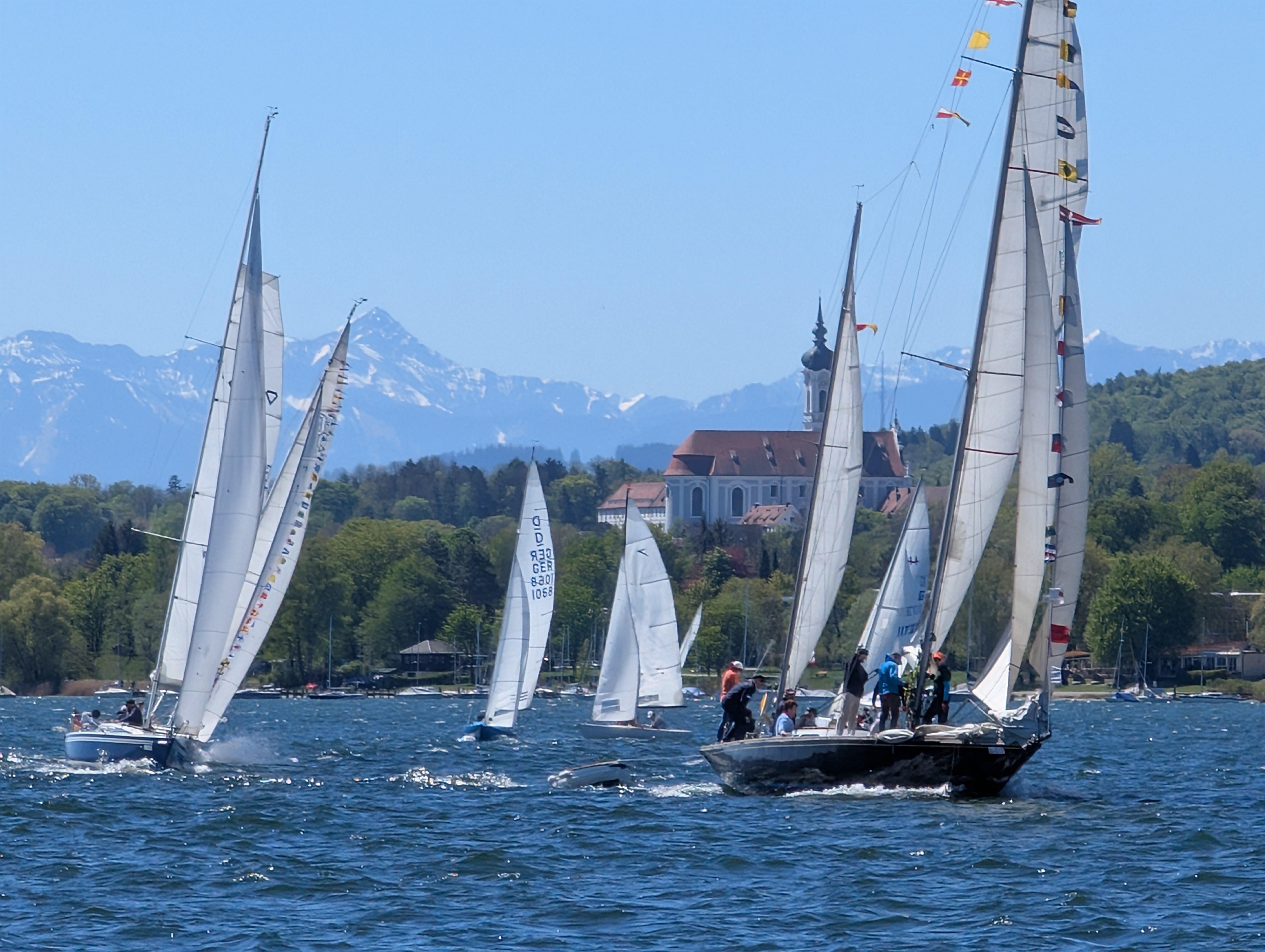 Einige Yachten segeln auf einem See. Die Sonne scheint, das Wasser glitzert. Im Hintergrund steht am Ufer eine Kirche, dahinter sind hohe Berge.