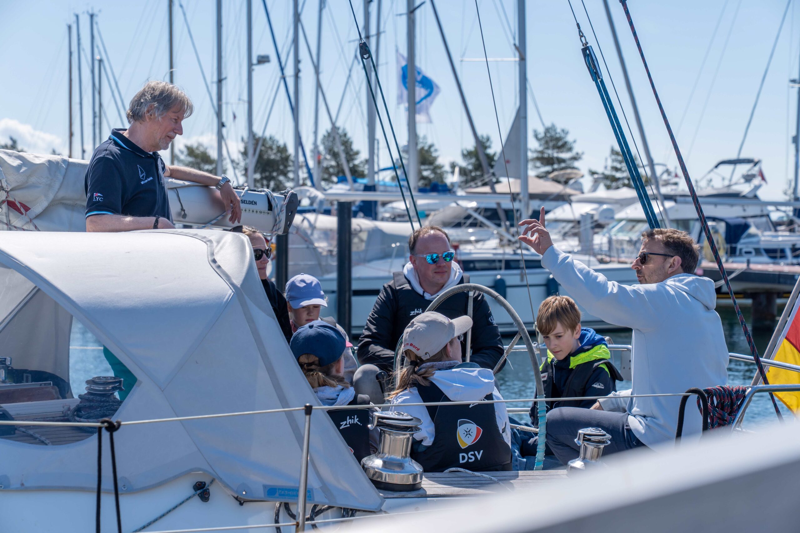 Ein Mann erklärt im Cockpit einer Yacht sitzenden Menschen wie Segeln funktioniert.