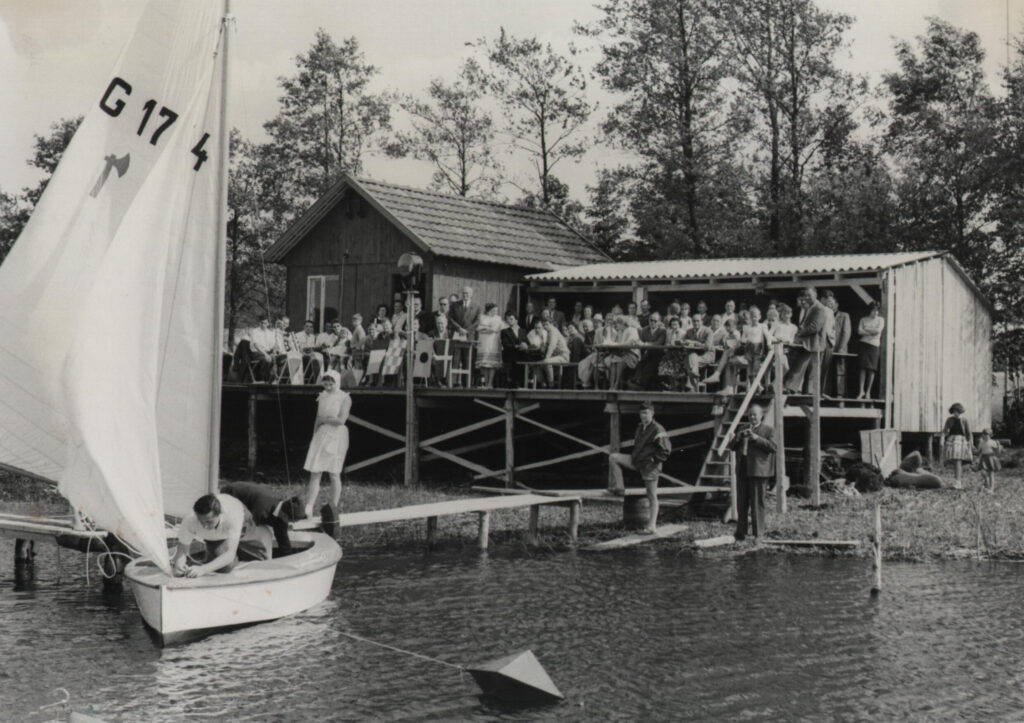 Das Foto ist ein historisches Schwarz-Weiß-Foto. Es zeigt Menschen auf einem Steg, die ein neues Segelboot beklatschen. Es ist eine Bootstaufe.