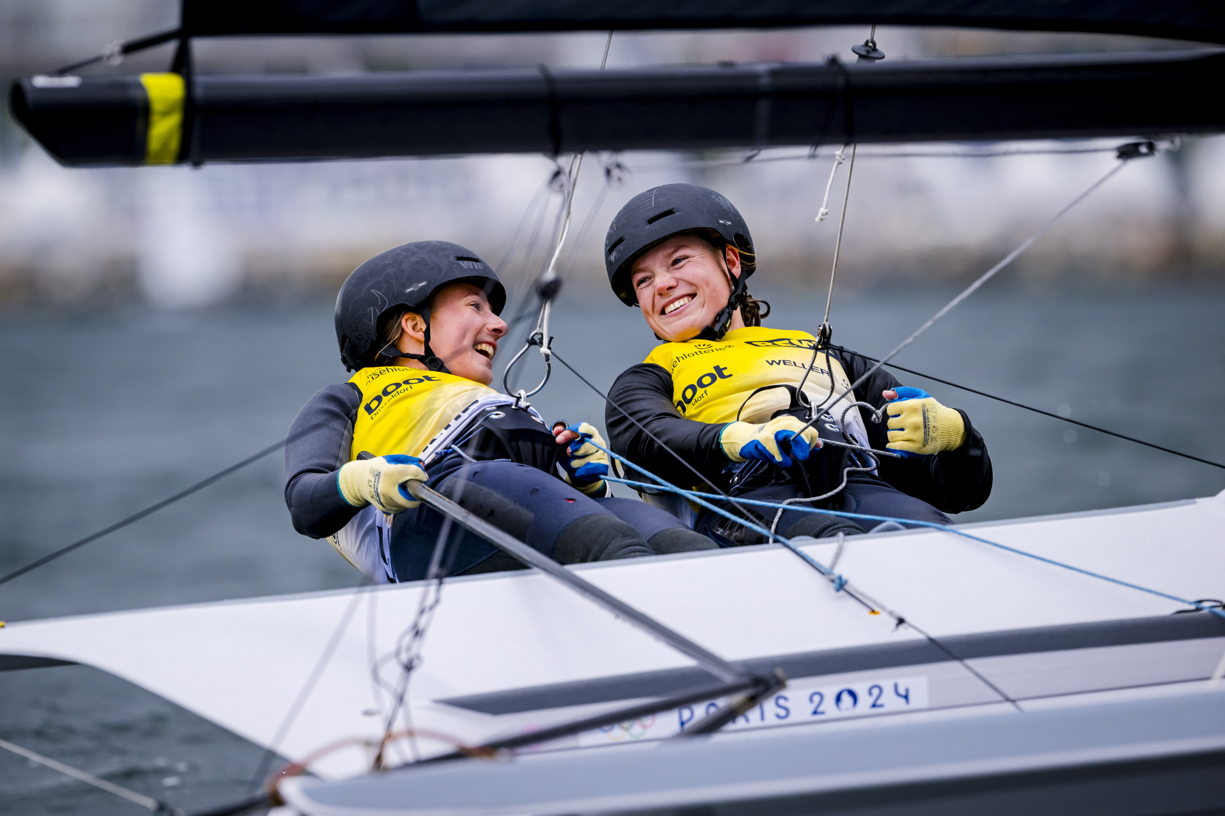 Zwei Frauen auf einer schnellen Skiffjolle mit schwarzen Helmen und gelben Trikots, sie lachen sich an und haben gute Laune.