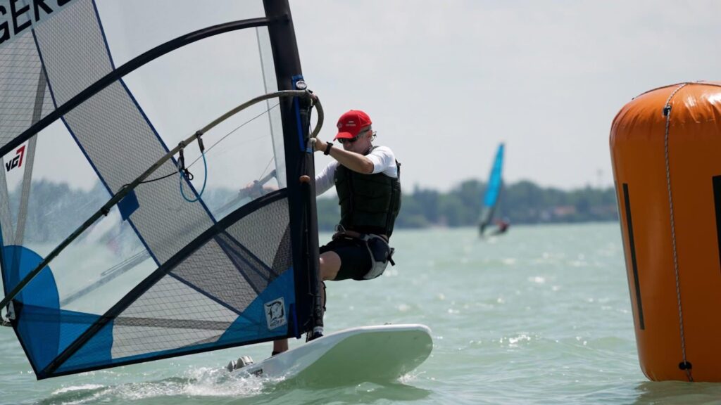 Ein Surfer rast über das Wasser. Er trägt eine rote Kappe und eine Sonnenbrille. Das Wasser glitzert.