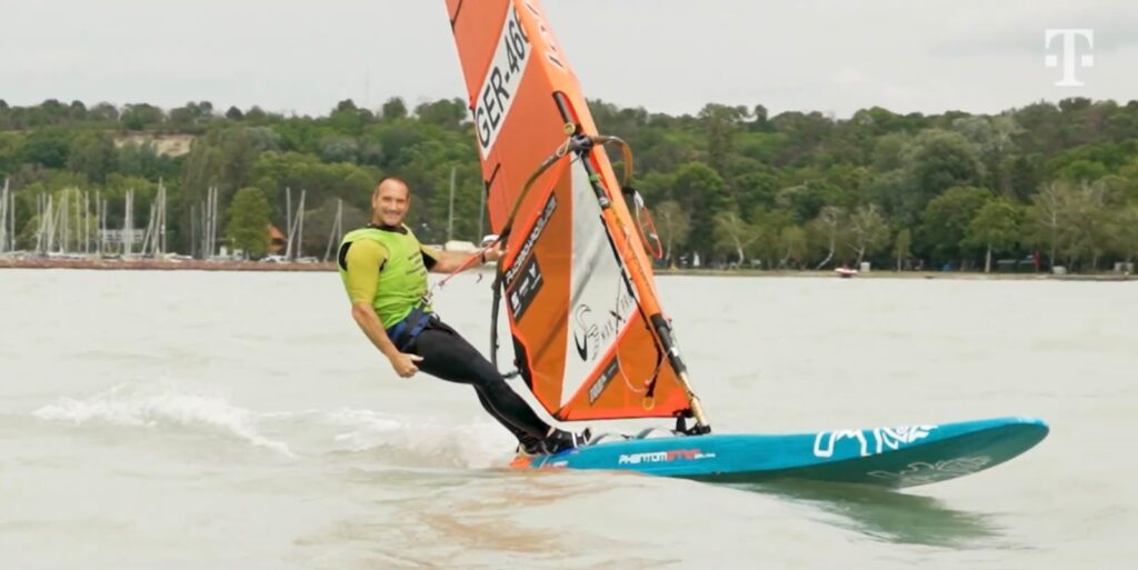 Ein Surfer surft über das Wasser und freut sich. Er hängt im Trapez, Das Segel ist orange, das Board türkis
