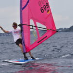 Ein Windsurfer surft über das Wasser, er streckt den Arm in die Höhe und freut sich. Das Segel ist pinkfarben