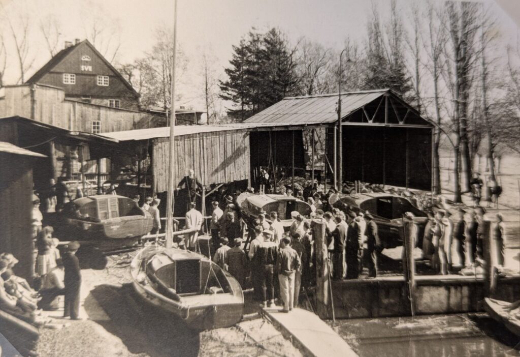 Das Schwarz-weiß Foto ist ein historisches Foto, Menschen stehen in einem Hafen und hören einem Mann zu, der eine Rede hält.