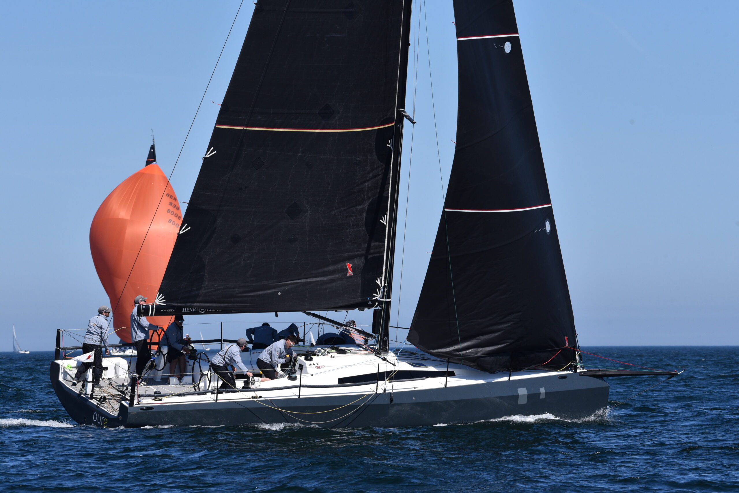 Eine schwarze Rennyacht vom Typ Mat 1220 von der Seite, im Hintergrund der rote Spinnaker einer anderen Yacht.