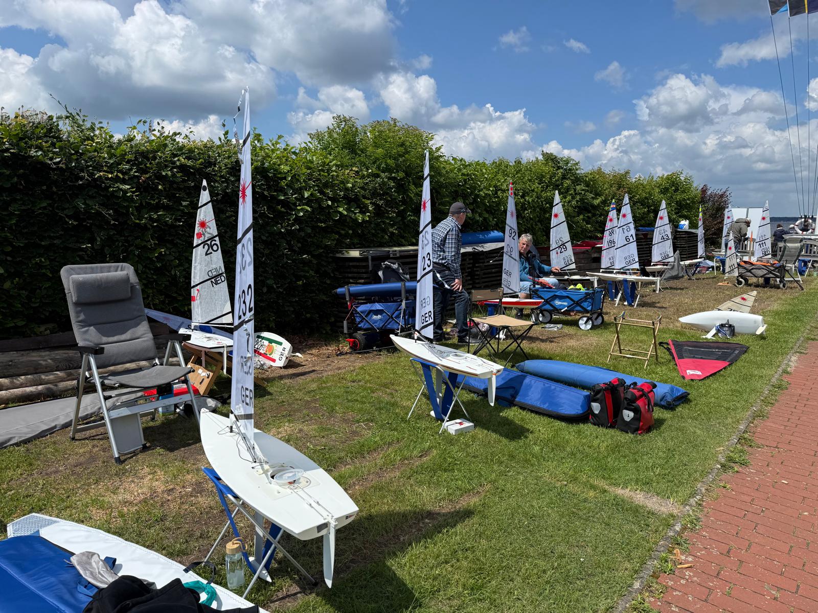 Kleine funkgesteuerte Yachten stehen an Land. die Fahrerinnen und Fahrer sitzen daneben. Der Himmel ist blau und wolkig.