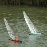 Zwei kleine Modellsegelboote segeln über das Wasser. Eines hat einen türkisfarbenen Rumpf, das andere eine schwarz-orangen