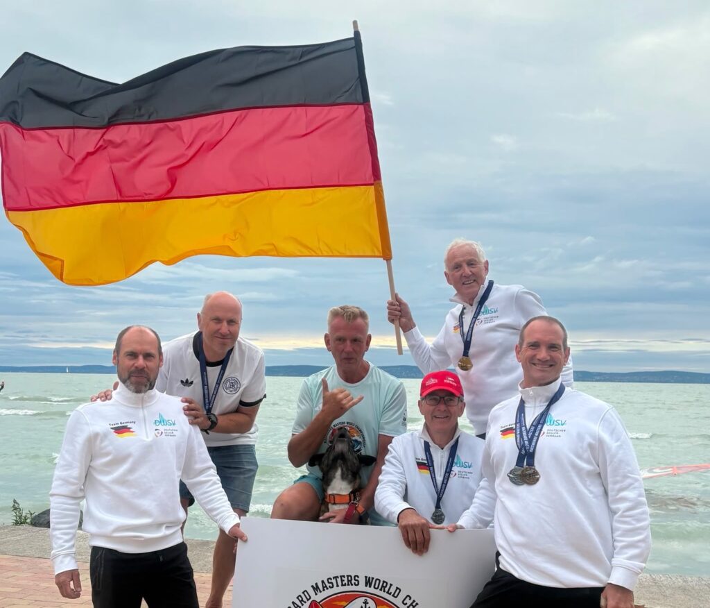Fünf Männer stehen am Strand zusammen, einige tragen Medaillen um den Hals, einer zusätzliche eine deutsche Fahne. Alle freuen sich.