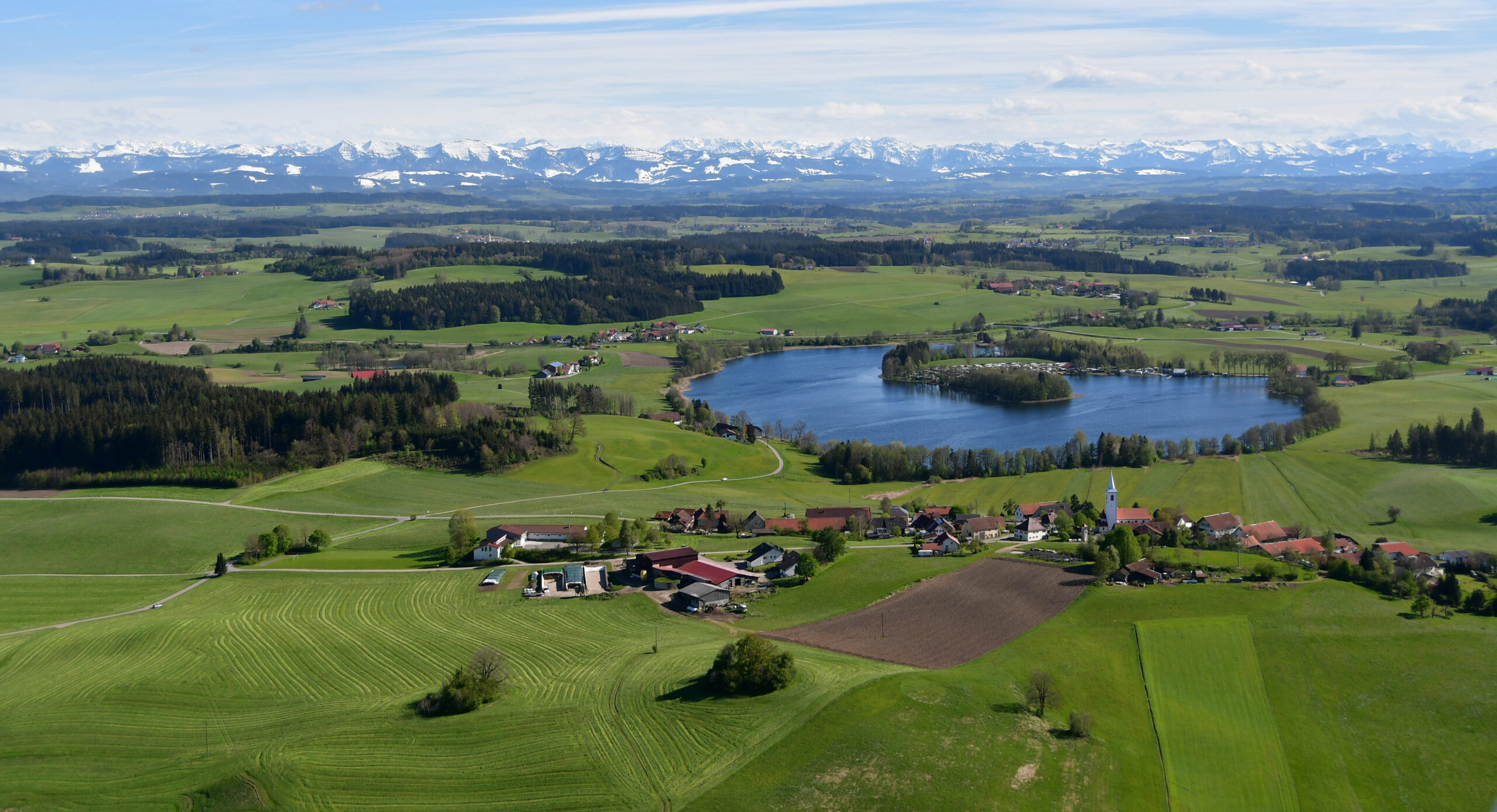 Die Luftaufnahme von oben zeigt einen kleinen Teich inmitten von Feldern, im Hintergrund sind Berge.