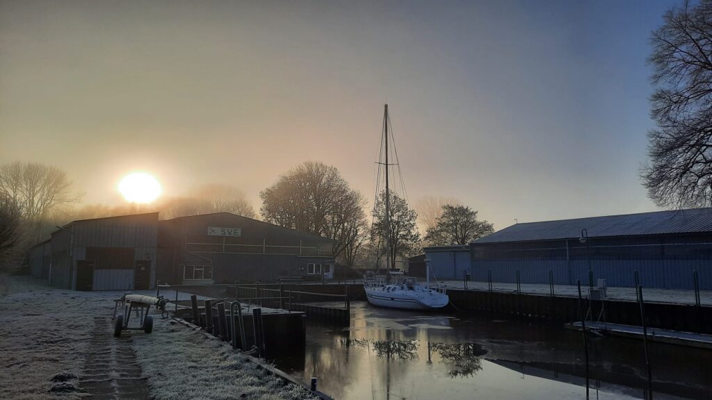 Hinter einer Bootshalle geht die Sonne auf, es ist neblig, im Hafenbecken davor liegt eine Yacht .