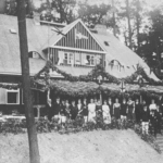 Das historische schwar-weiß Foto zeigt ein haus, es ist das Clubhaus. Vor dem Haus stehen Fraune und Männer und freuen sich.