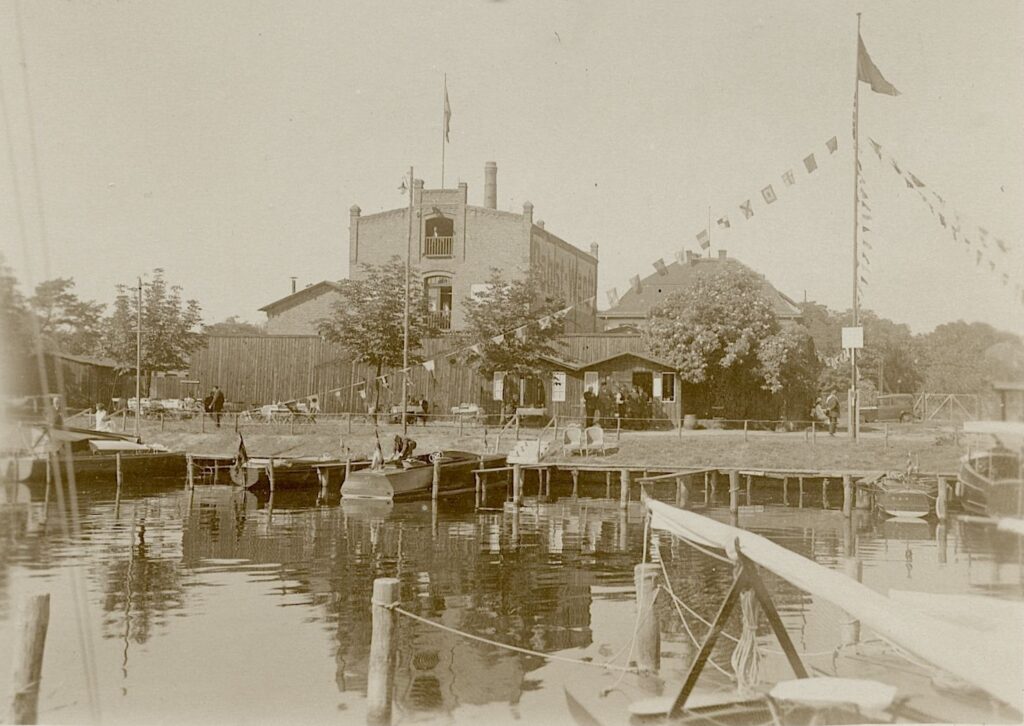 Das historische schwar-weiß Foto zeugt ein Gebäüde, im Vordergrund liegen Segelboote am Ufer.