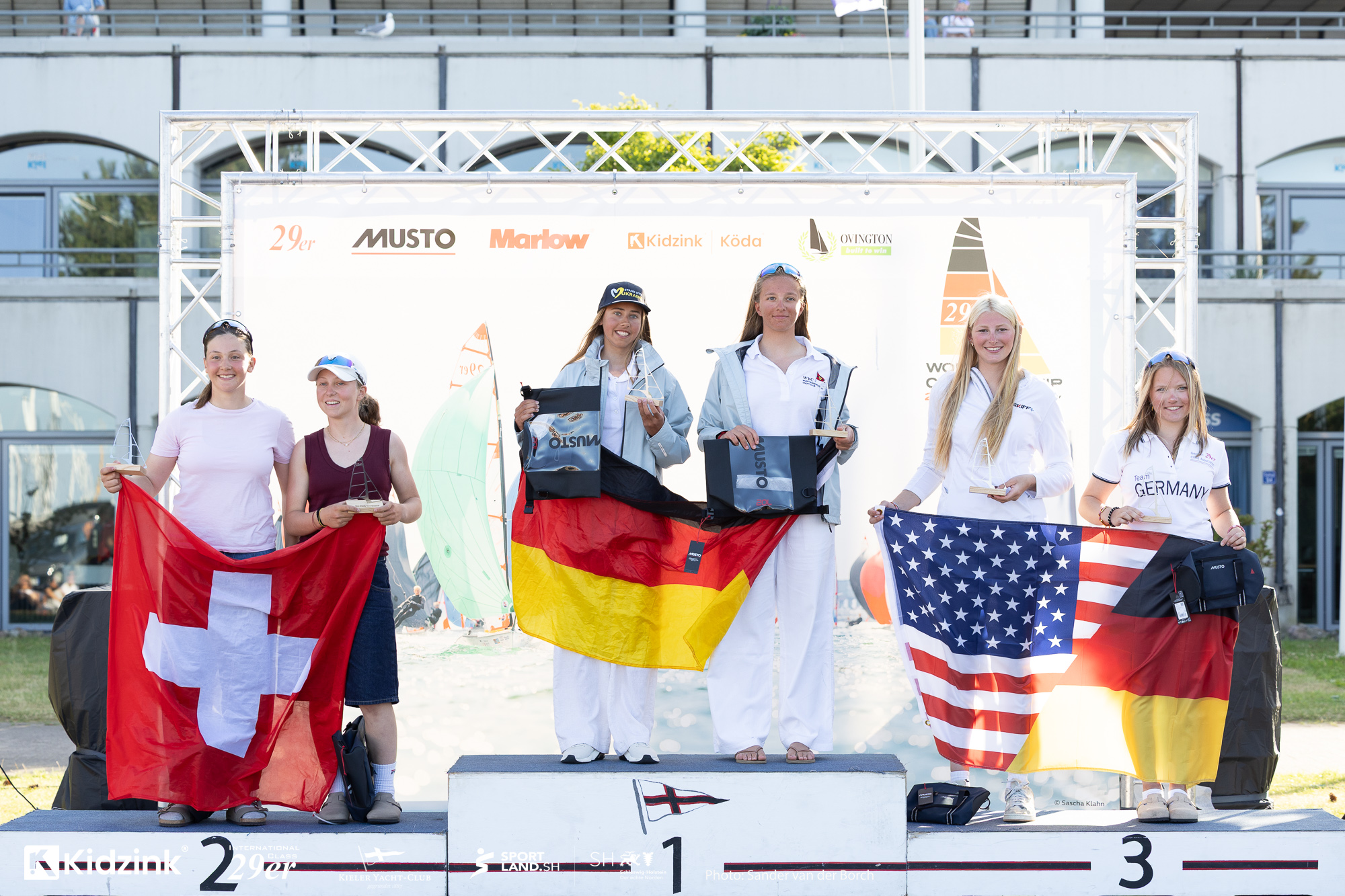 Sechs Mädchen stehen auf dem Siegerpodest nach einer Regatta, sie zeigen die deutsche, schweizer und US-amerikanische Flagge.