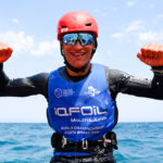 Ein Mann mit einem blauen Trikot, verspiegelter Brille und einem roten Helm jubelt auf dem Wasser