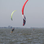 Auf einem See kiten Kiter und Kiterinnen. Am Himmel schweben ein rot-Weißer und ein Blau-roter Kite.