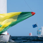 Ein grün-gelber Spinnaker von einer Jolle vom Typ Flying Dutchman, dahinter rechts eine blaue Zielflagge.