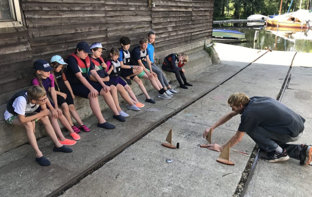 Kinder sitzen auf einer Bank an einem Holzhaus, vor ihnen demonstriert ein Trainer mit kleinen Bootsmodellen auf dem Boden die Vorfahrtsregeln beim Segeln.