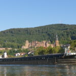 Segeln auf einem Fluss, vorne ein kleines Segelboot, dahinter eine große flache Schute, am Ufer steht ein historisches Schloss