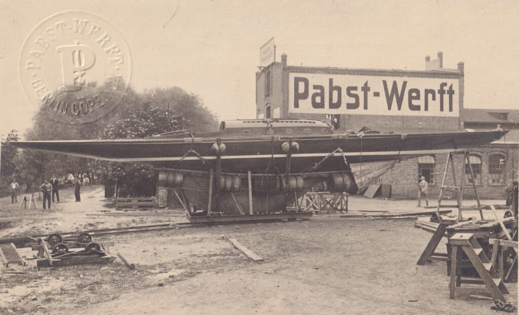 Das historische schwarz-weiß Foto zeugt im Hintergrund ein Haus, im Vordergrund steht der Rumpf einer Holzyacht auf einem Gedtell. Das Foto ist auf einer Werft entstanden