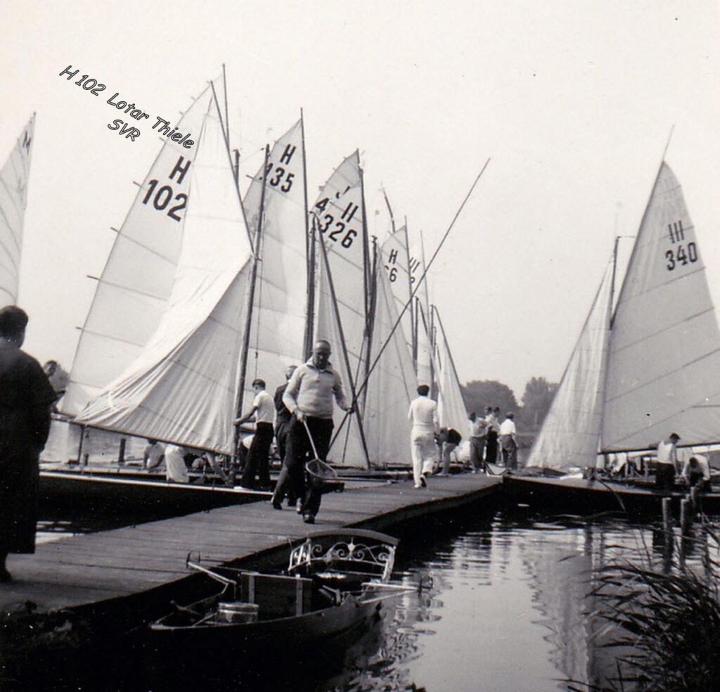 Das historische Foto ist schwarz-weiß. Boote liegen mit gesetzten Segeln an einem Steg.