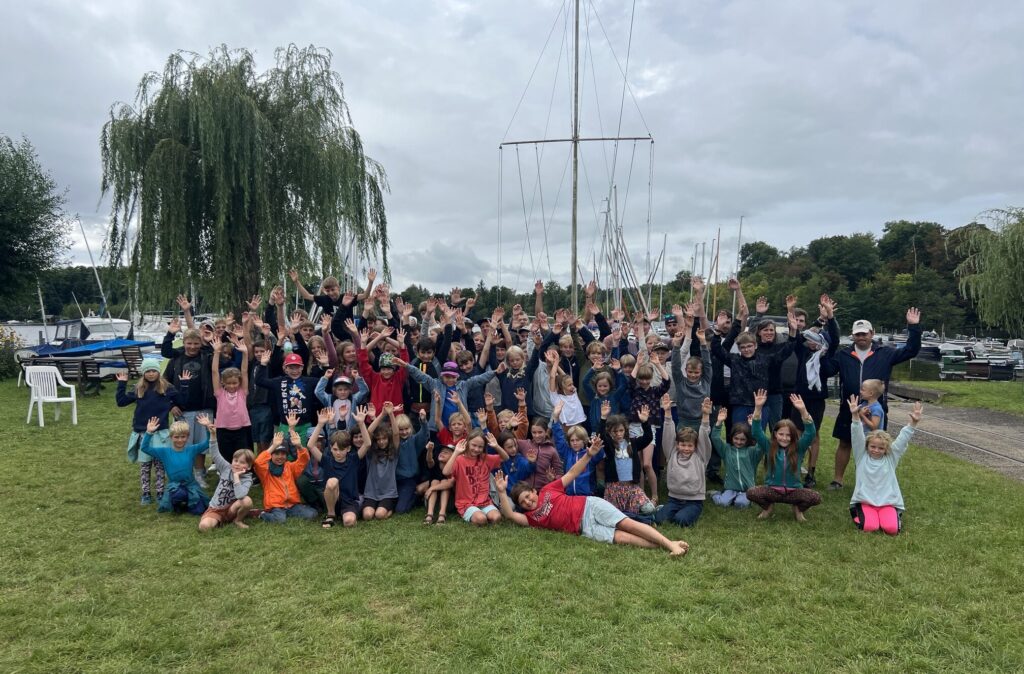 Vile Kinder haben sich zu einem Gruppenbild auf einer Wiese in einem Yachthafen versammelt. Im Hintergrund: Segelmasten