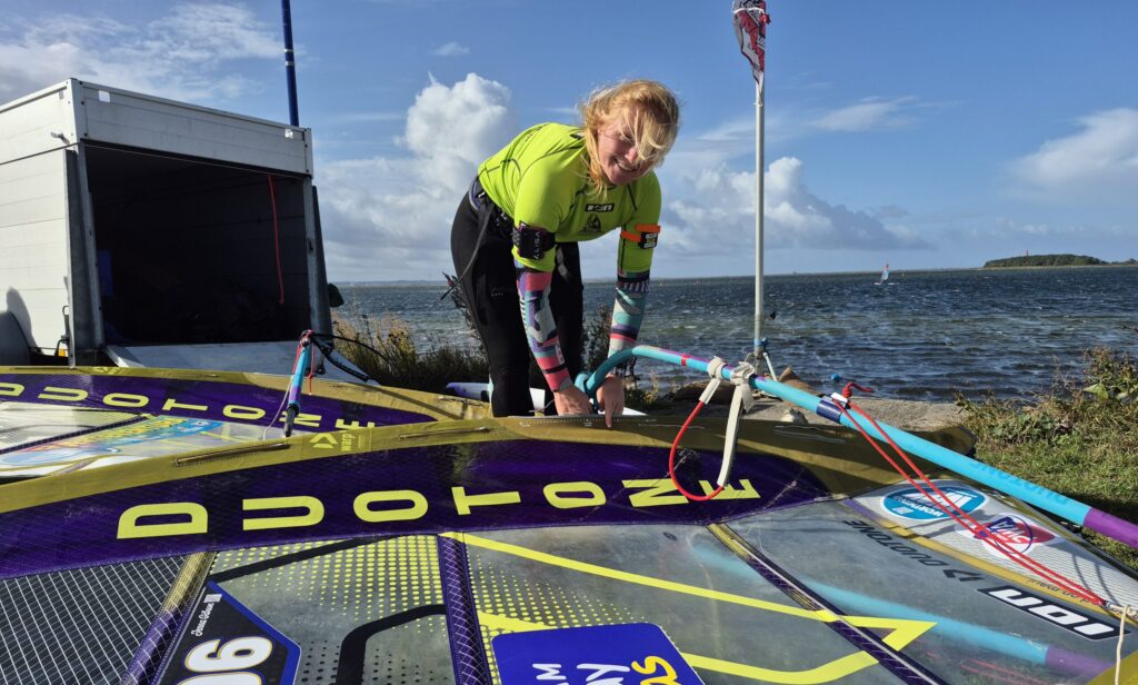 Eine Windsurferin trimmt ihr Rigg, im Hintergrund die Orther Bucht auf Fehmarn.