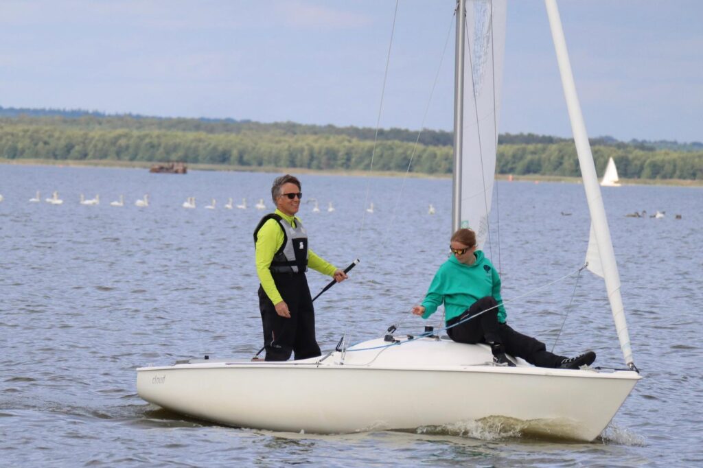 Ein Mann in einem gelben Pullover mit schwarzer Hose und Schwimmweste auf einem Conger, vorne im grünen Pullover ein junges Mädchen.