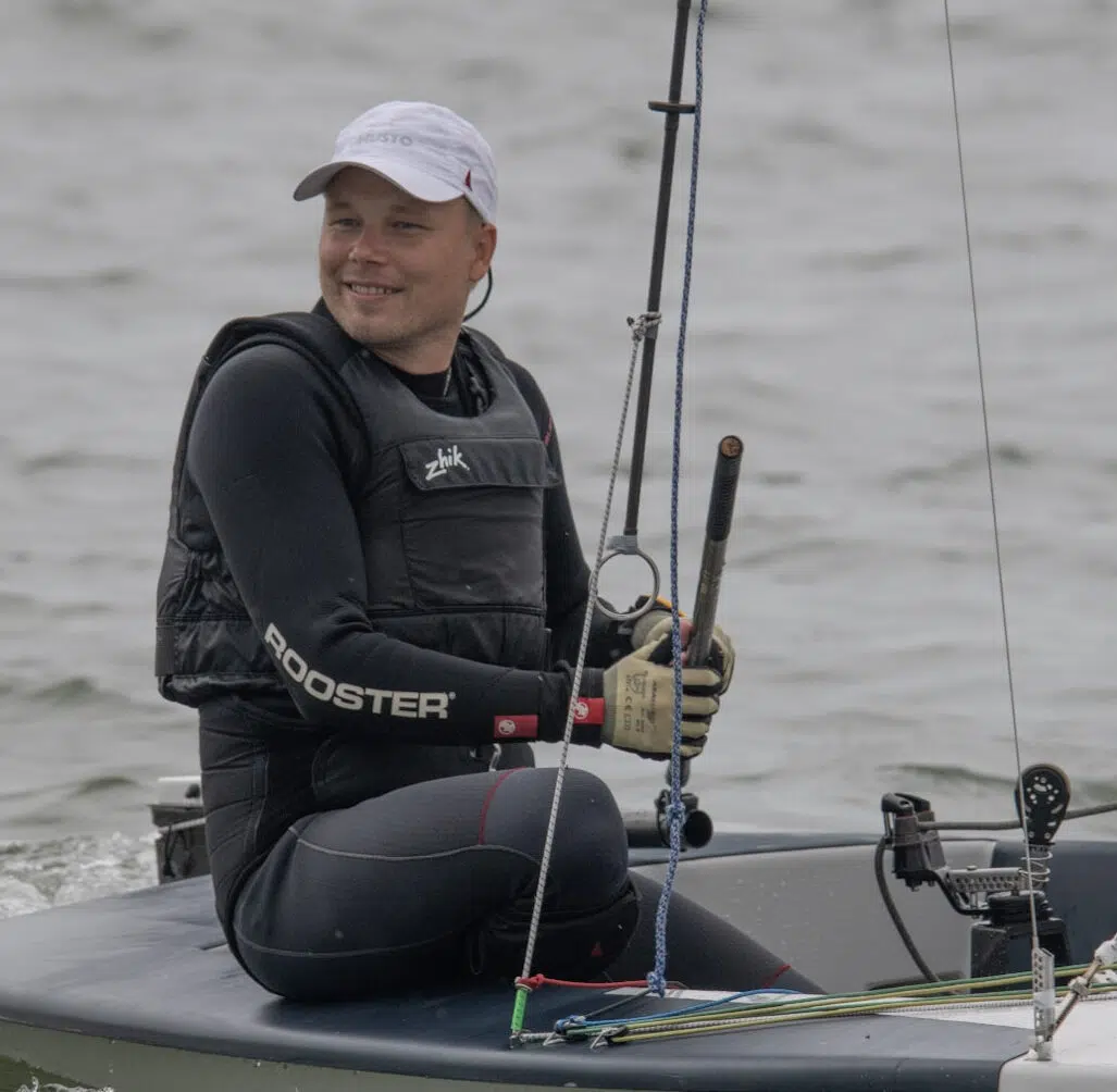 Contender-Segler Max Billerbeck auf seiner Jolle sitzend, er trägt einen schwarzen Neoprenanzug und ein weißes Cap und hat den Pinnenausleger in der Hand.