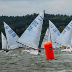 Mehrere Jollen vom Typ Finn Dinghy segeln mit Wind von hinten zu einer roten Tonne