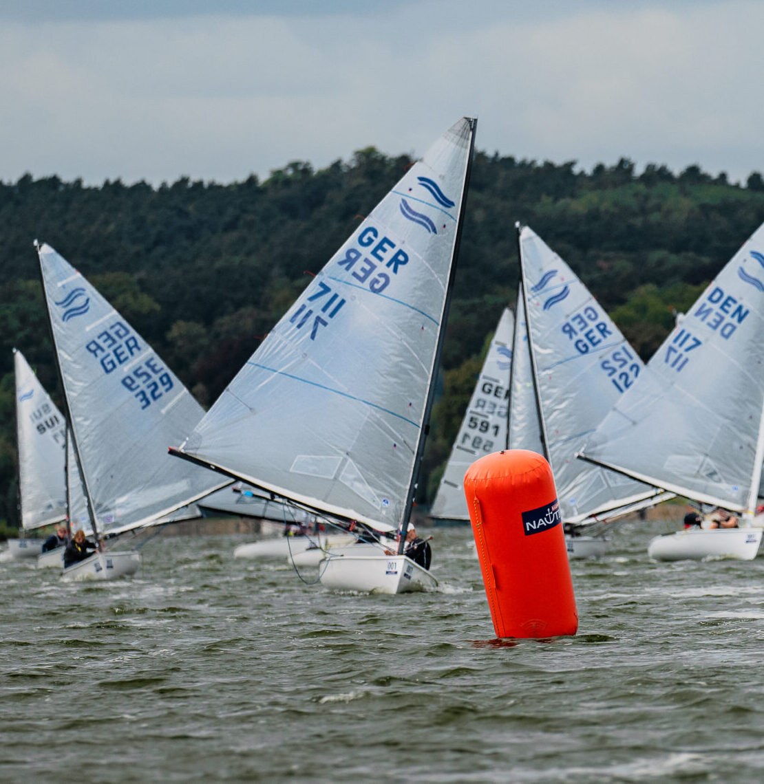 Mehrere Jollen vom Typ Finn Dinghy segeln mit Wind von hinten zu einer roten Tonne