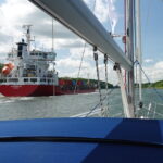 Eine Segelyacht fährt durch den Nord-Ostsee-Kanal hinter einem roten Containerschiff.
