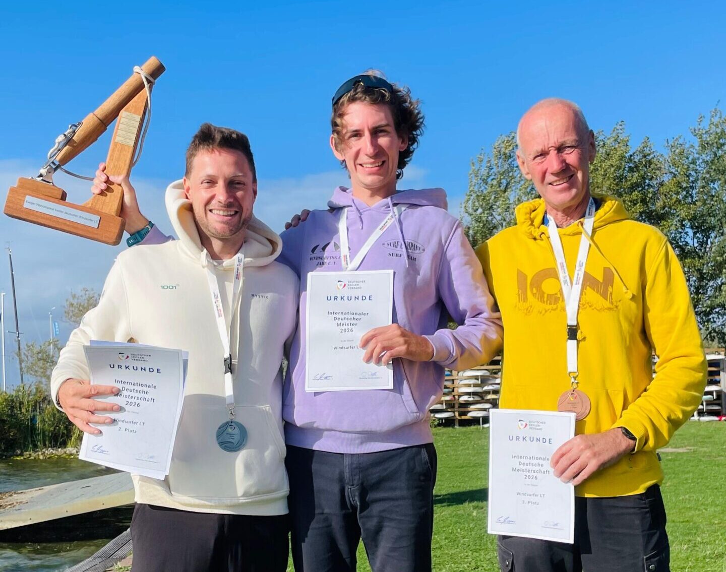 Drei Männer mit Hoodies und Urkunden in der Hand, der mittlere hält einen hölzernen Preis in der Hand, der einen Teil eines Surfmastes darstellt